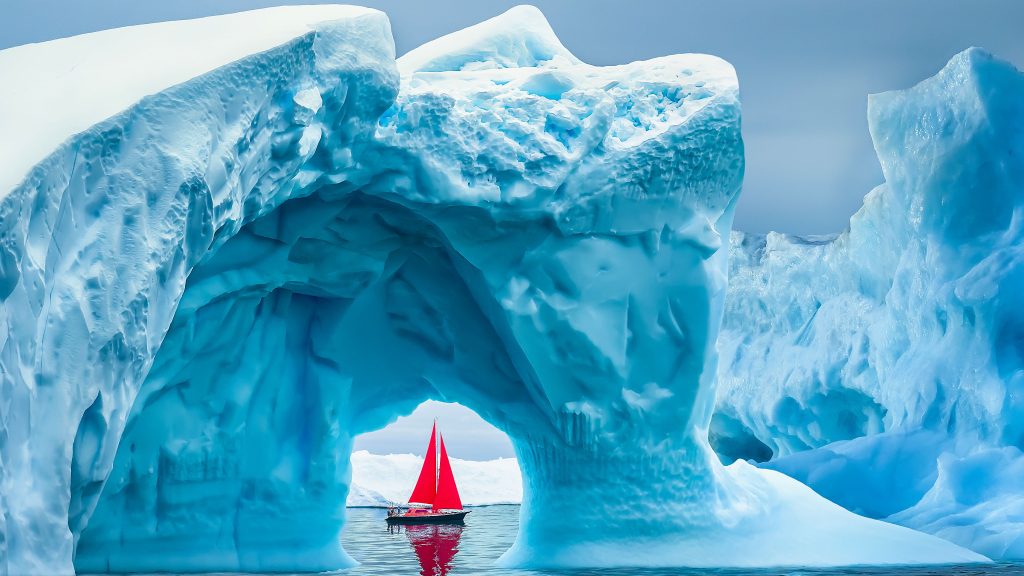 Red sailboat sailing under iceberg arch, Ilulissat Icefjord, Disko Bay, Greenland