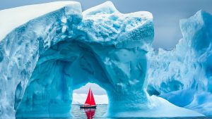 Read more about the article Red sailboat sailing under iceberg arch, Ilulissat Icefjord, Disko Bay, Greenland